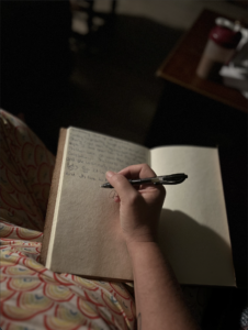 A hand writing with a black pen in an open notebook resting on the writer's lap.